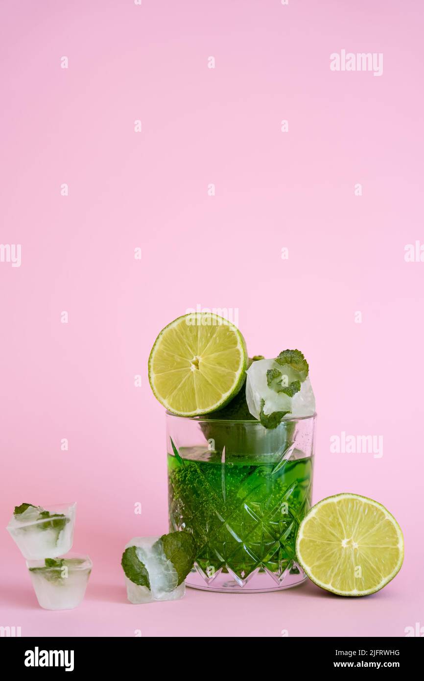 green and fresh limes in faceted glass with sparkling alcohol drink near ice cubes with peppermint on pink Stock Photo