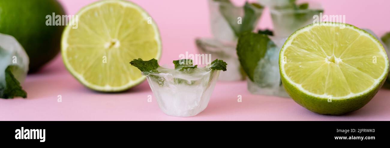green and fresh limes near blurred ice cubes with peppermint on pink, banner Stock Photo