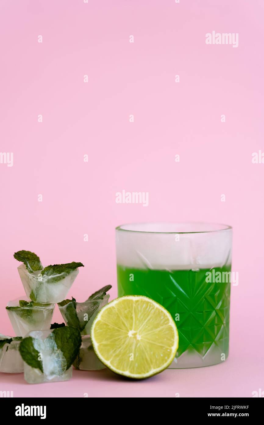 green and fresh lime in faceted glass with alcohol drink near ice cubes with peppermint on pink Stock Photo