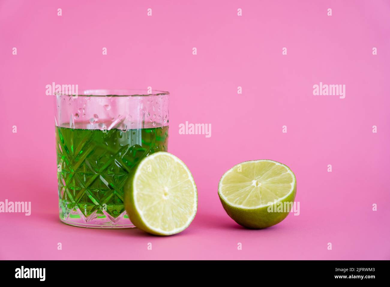 green alcohol drink in faceted glass with water drops near limes on pink Stock Photo