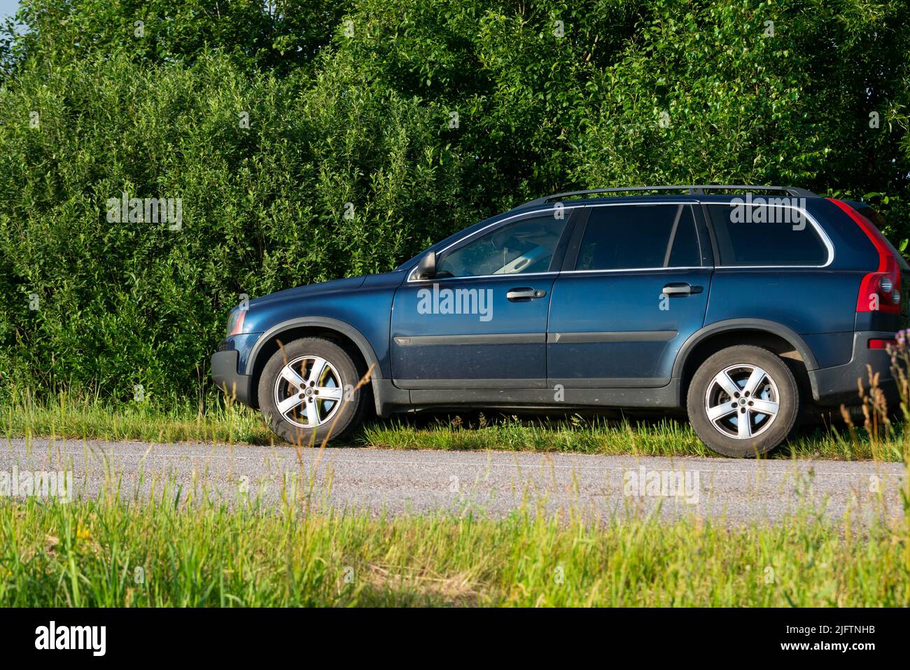 SUV on the side of a country road next to green bushes in green grass illuminated by warm summer sunset light. Stock Photo