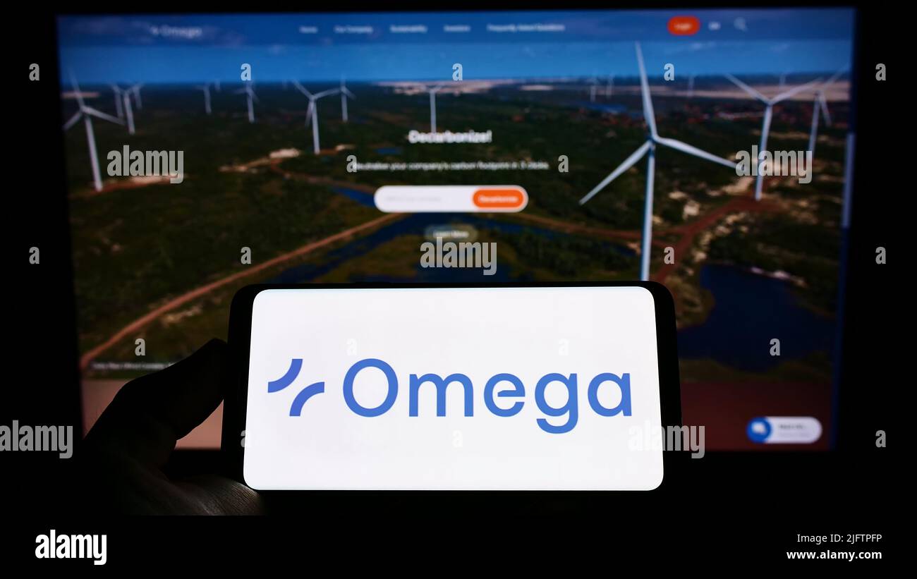 Person holding cellphone with logo of Brazilian renewable energy company Omega Energia on screen in front of webpage. Focus on phone display. Stock Photo