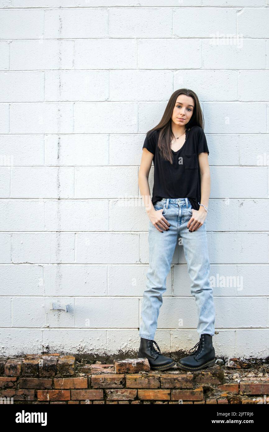A teenage girl with long hair wearing black, denim and combat boots leaning against an old cinder block wall and looking down with attitude Stock Photo