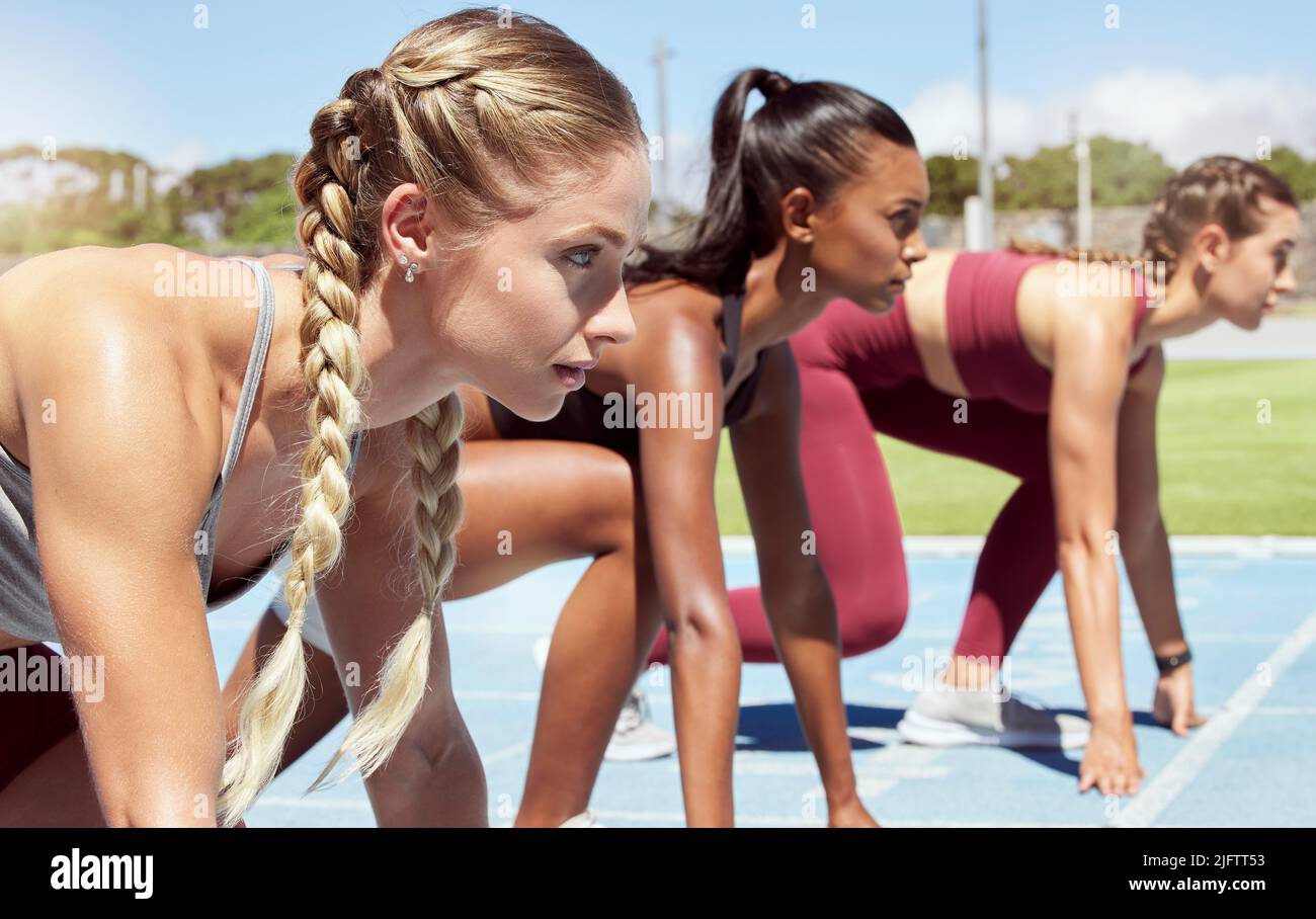 Three female athletes at the starting line in a track race competition at the stadium. Young sporty women in a race waiting and ready to run. Diverse Stock Photo