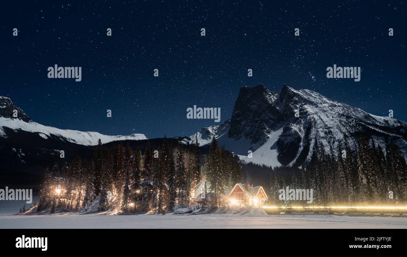 Iluminated alpine huts amongst the trees under the mountains and sky full of stars, winter, Canada Stock Photo