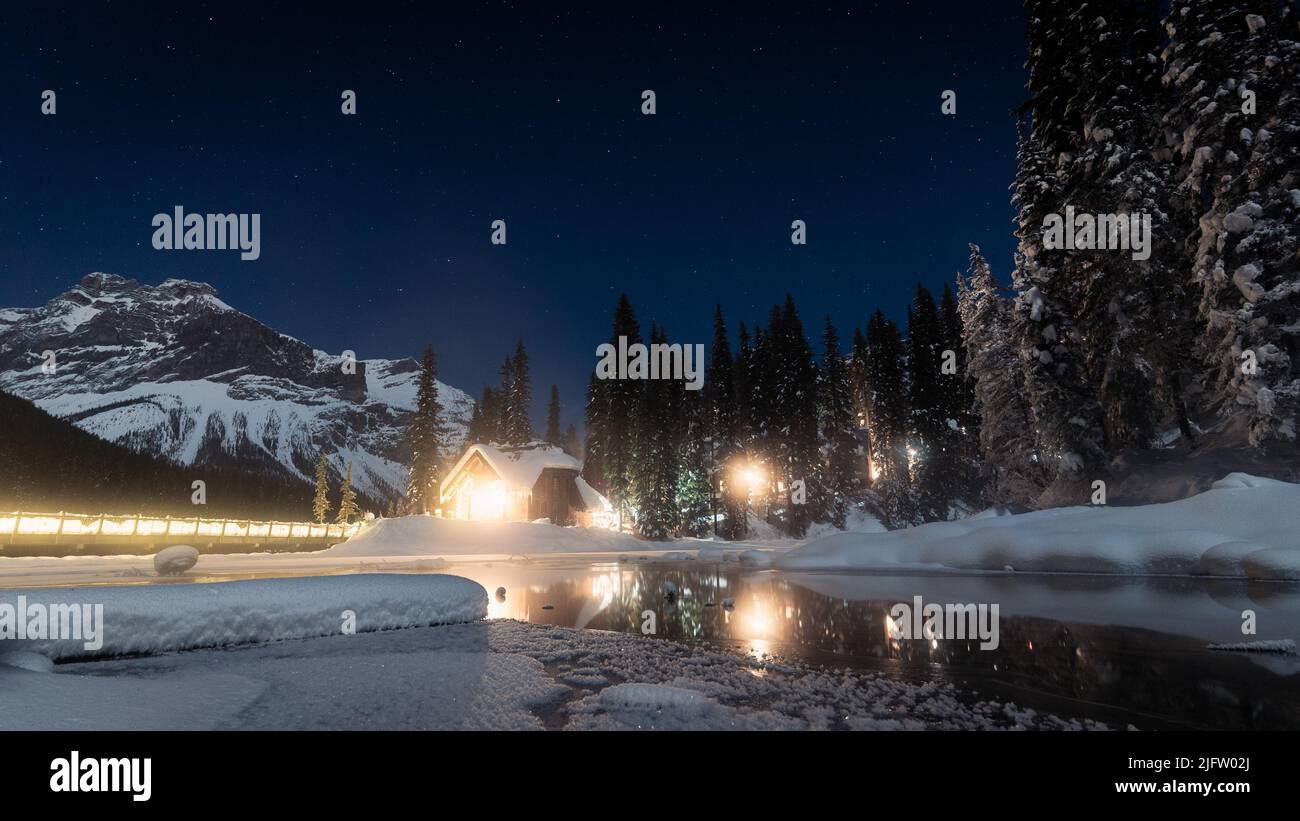 Small hut on the bank of frozen lake surrounded by mountains with stars above, Yoho N. Park, Canada Stock Photo
