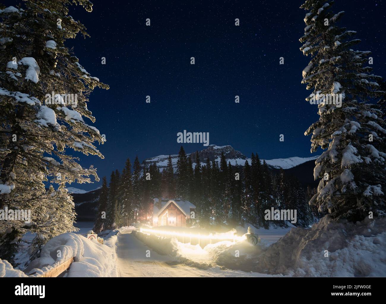 Small hut in a winter forest surrounded by trees, mountains with bright stars above, Yoho NP, Canada Stock Photo