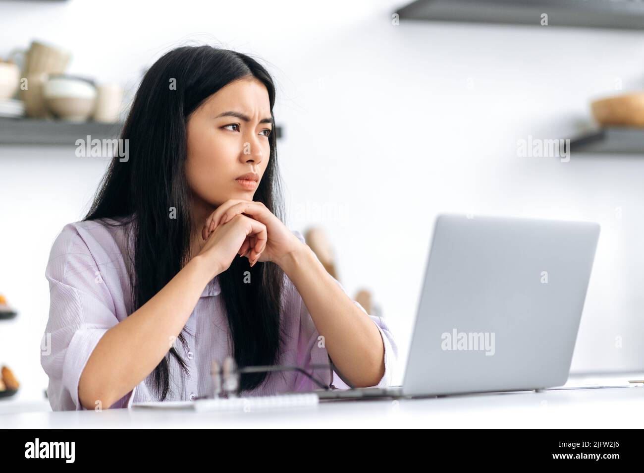 Sad unhappy Asian millennial girl, student or freelancer, sits at the table with a laptop, upset by a bad message, dissatisfied with the result of the project, did not receive approval, in a bad mood Stock Photo