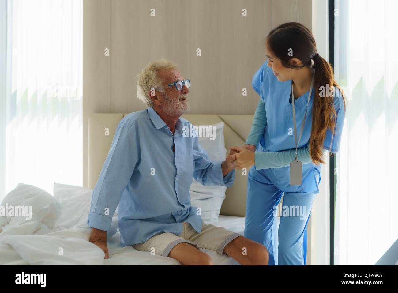 Asian nurse standing on a home bed next to an older man helping hands, care. Elderly patient care and health lifestyle, medical concept. Stock Photo