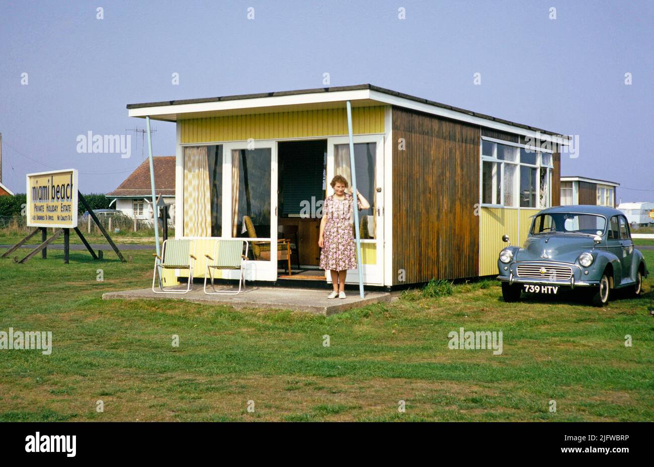 Woman standing in doorway of holiday chalet with Morris Minor car parked, Miami Beach private holiday estate, Sutton on Sea, Lincolnshire, England, UK, 1960s Stock Photo