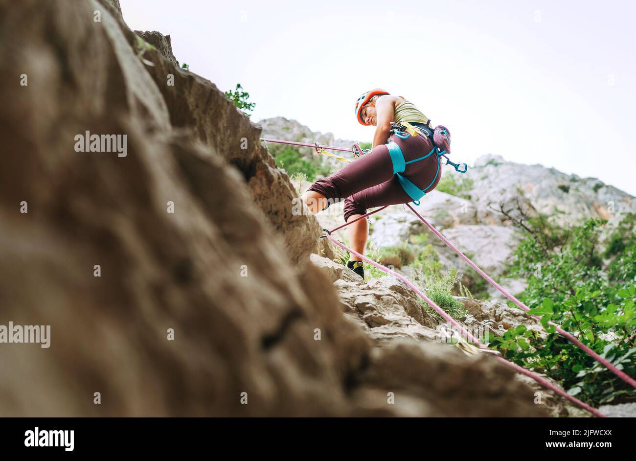 Active climber woman in protective helmet abseiling from cliff rock wall using rope with belay device and climbing harness. Active extreme sports time Stock Photo