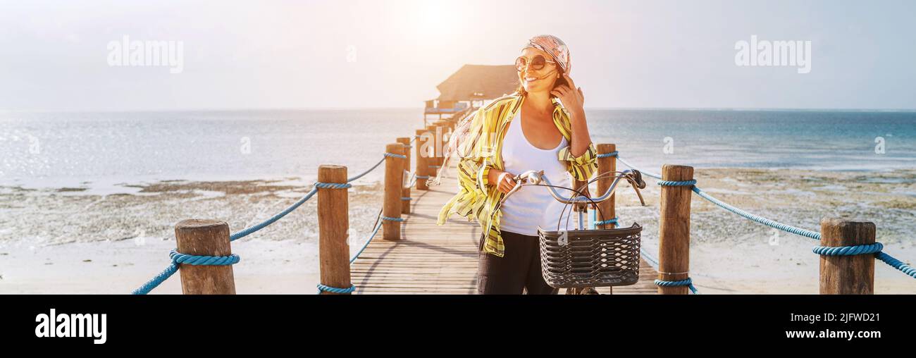Portrait of a smiling woman dressed in light summer clothes, sunglasses with bicycle on the wooden sea pier on the sandy Zanzibar beach.Careless vacat Stock Photo