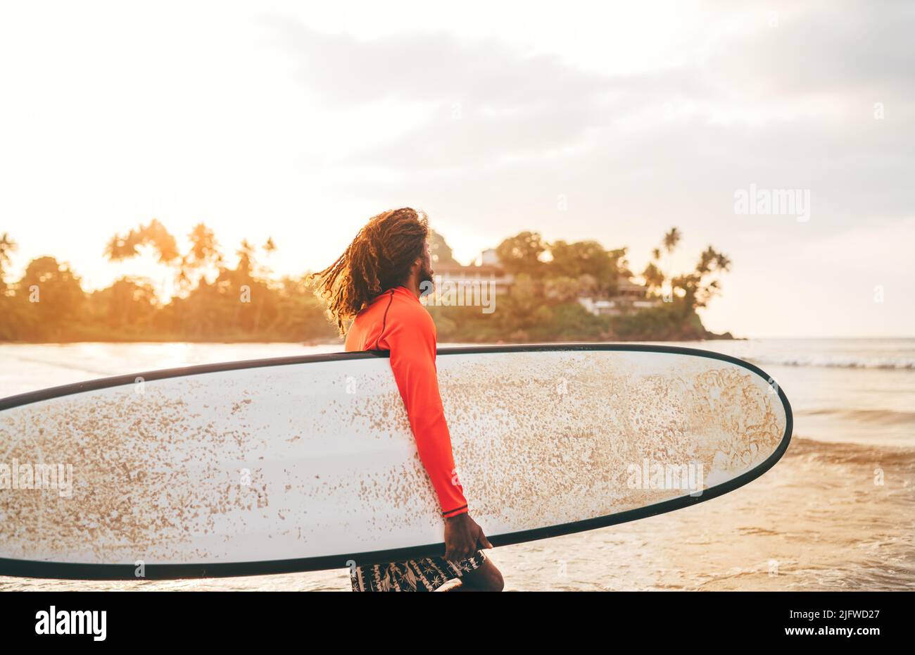 Black long-haired teen boy with a waxed surfboard ready for surfing with sunset backlight. He walking into Indian ocean waves. Extreme water sports an Stock Photo
