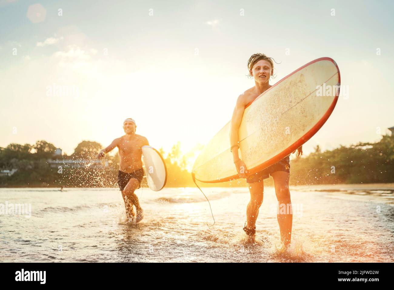 Young teen boy with a surfboard running with his father by ocean sandy beach after surfing. They are smiling and enjoying a beautiful sunset light. Fa Stock Photo