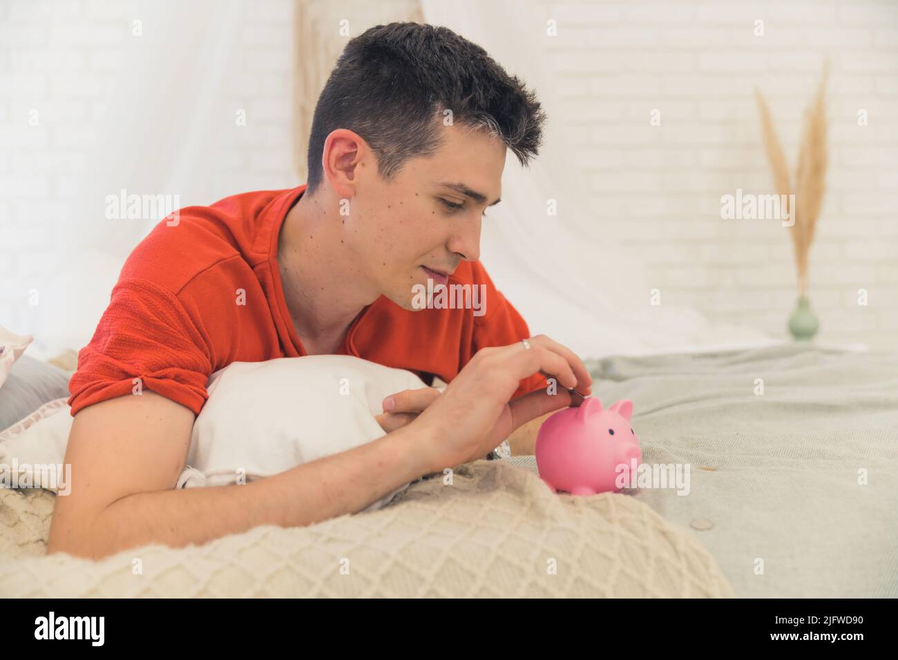 Responsible young caucasian dad worried about economic situation, laying down on his bed and putting a coin into a pink piggybank. Symbolic shot. High quality photo Stock Photo