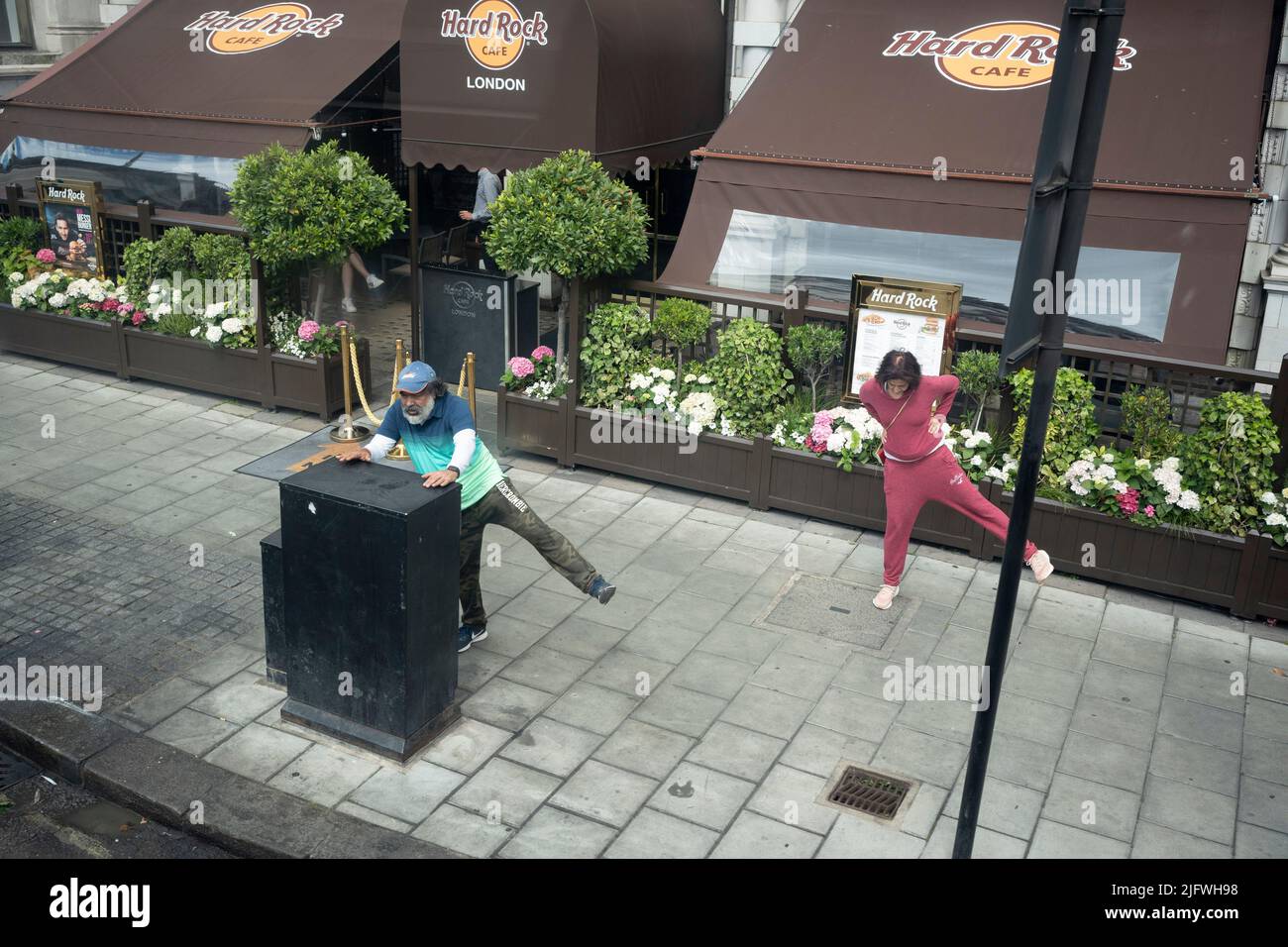 Seen from an aerial view of a London bus, are two middle-aged people who are together performing leg stretches outside the Hard Rock Cafe on Piccadilly in the capital, on 5th July 2022, in London, England. Stock Photo