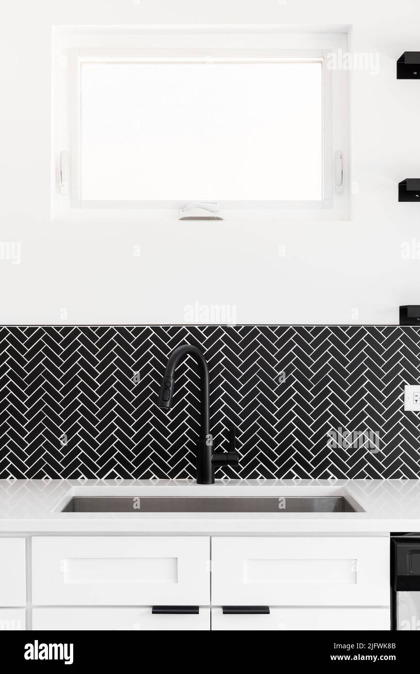A kitchen sink detail shot with a black faucet, black marble herringbone backsplash, and a white granite countertop. Stock Photo