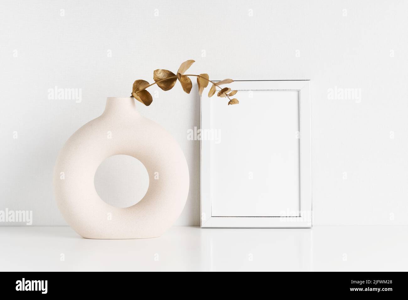 White frame mockup and round ceramic vase with a golden-colored eucalyptus branch on a white table by the wall. Copy space for text Stock Photo