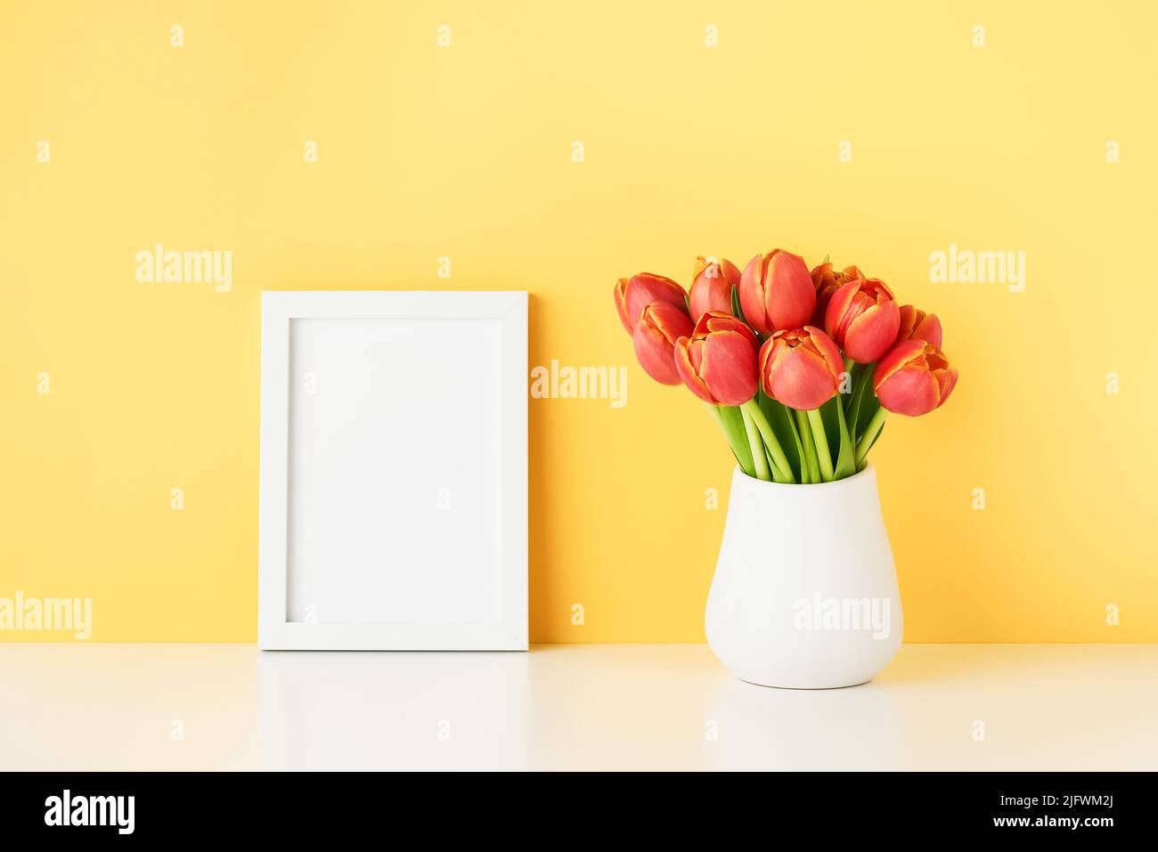 White frame mockup and red tulips in a white vase on a white table by the yellow wall. Copy space for text Stock Photo