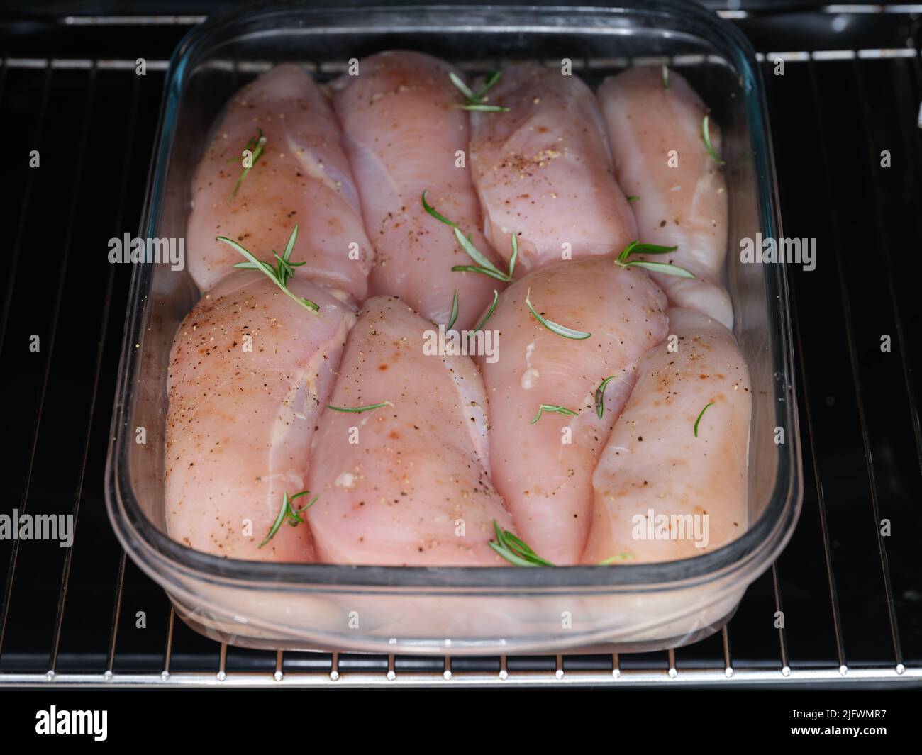A glass baking tray with raw chicken breasts with rosemary leaves on them in it in an oven. Close up. Stock Photo