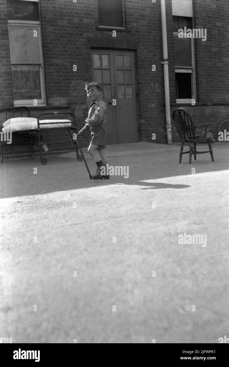 1942, historical, a little boy, a cub scout with special shoes on and holding a walking stick standing in the courty outside the wards at the Queen Mary's Hospital for Children at Carshalton, near London, England, UK. Built in 1908 as the Southern Hospital, it became a children's infirmary in 1909 and renamed after a visit by Queen Mary in 1915. It was the most heavily bombed hospital in the greater London area, with the first attack in 1940. The new threats posed by the VI and V2 rockets, saw total evacuation take place in July 1944 with children taken to other locations in England and Wales. Stock Photo