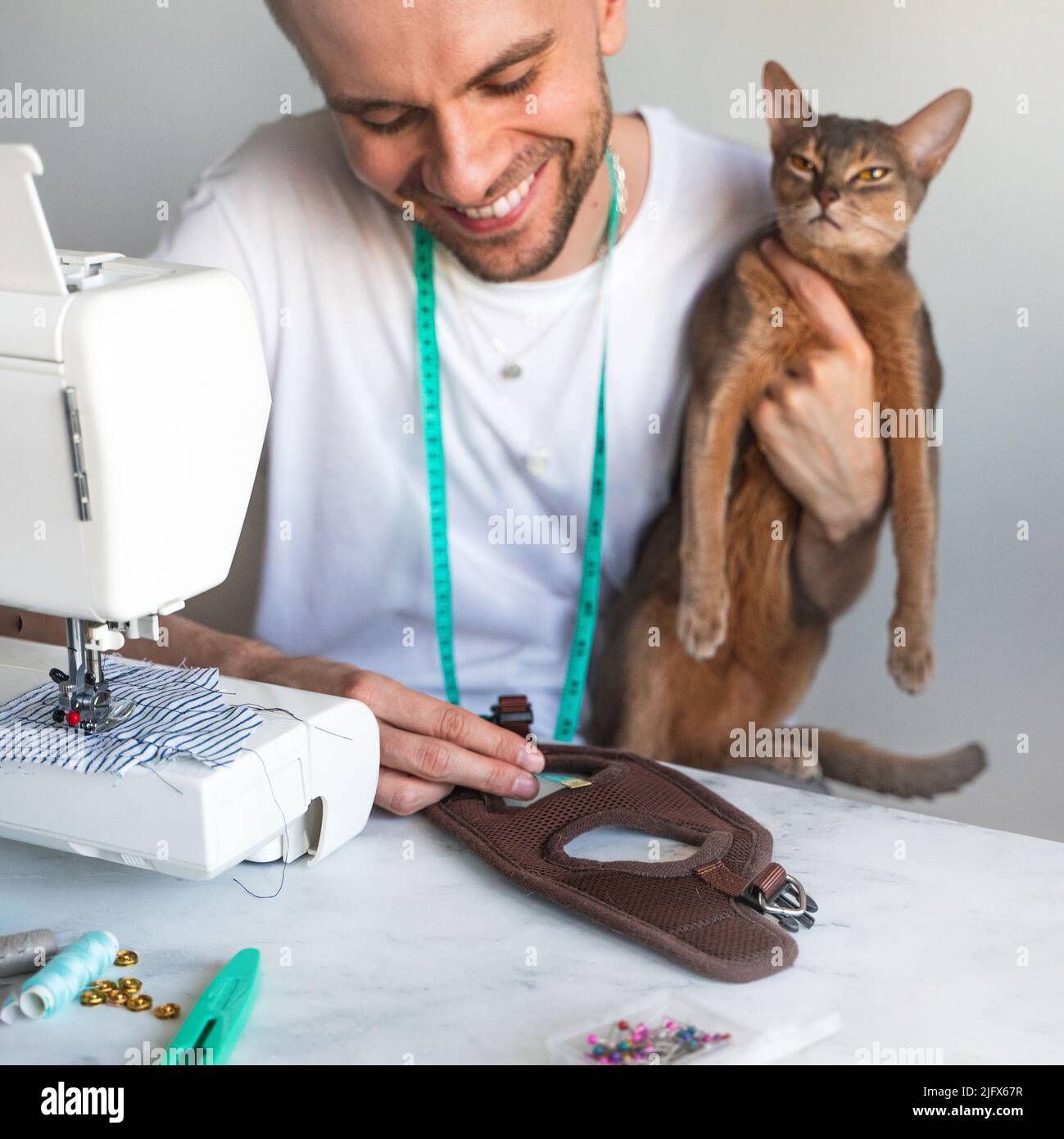 Sewing clothes for pets. Tailoring clothes for cats. A laughing tailor in  white t-shirt is sewing clothes for his abyssinian cat. DIY business  concept Stock Photo - Alamy, image size:1299x1390