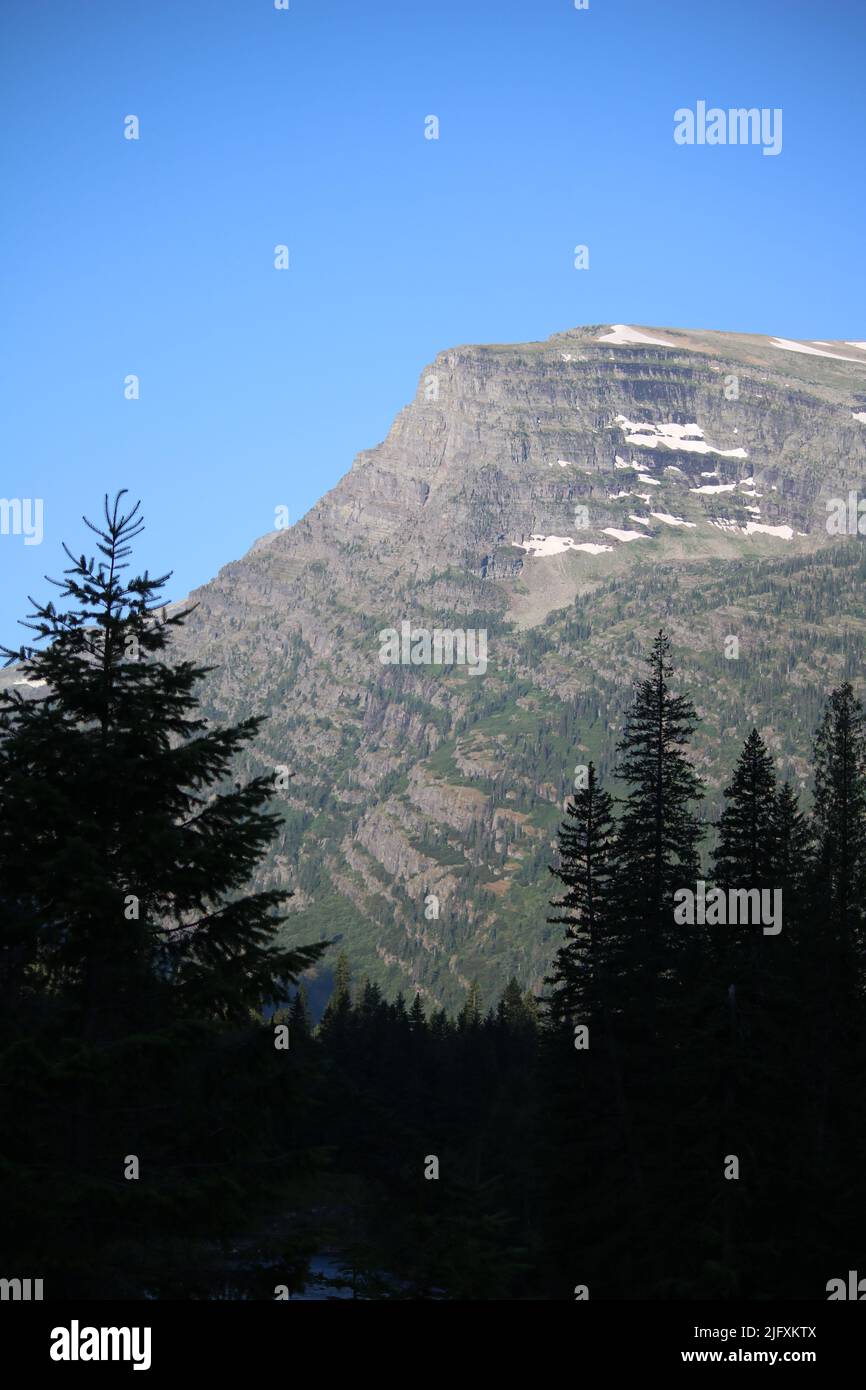 Glacier National Park Montana USA - Going to the Sun Rd, brilliant blue sky over majestic mountain peak with melting snow, pine forest, McDonald Creek Stock Photo