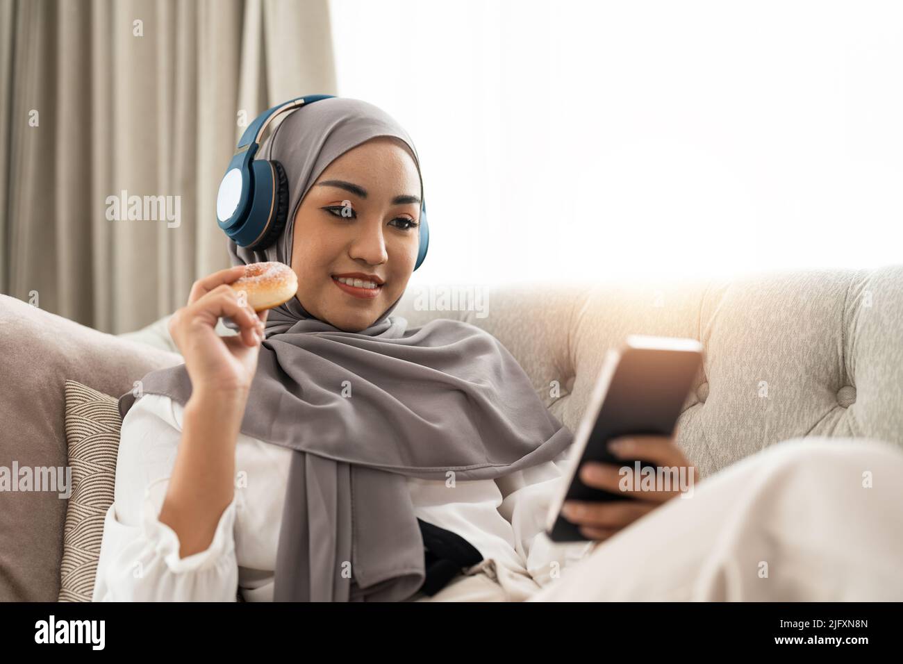Beautiful Asian muslim woman using mobile phone while sitting on sofa, girl doing chat text message on smartphone and smiling Stock Photo