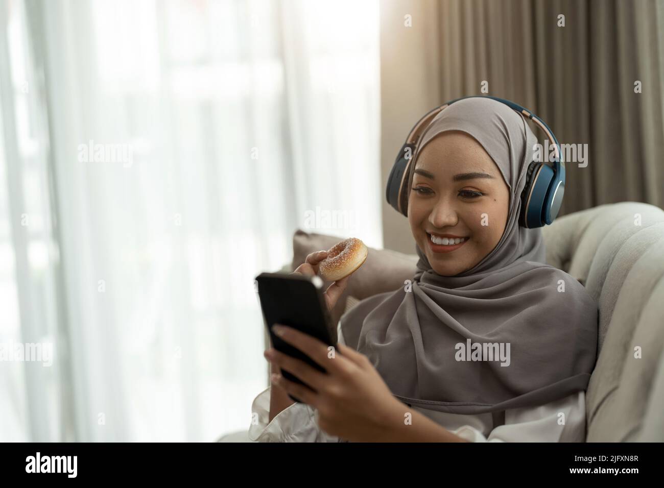 Beautiful Asian muslim woman using mobile phone while sitting on sofa, girl doing chat text message on smartphone and smiling Stock Photo