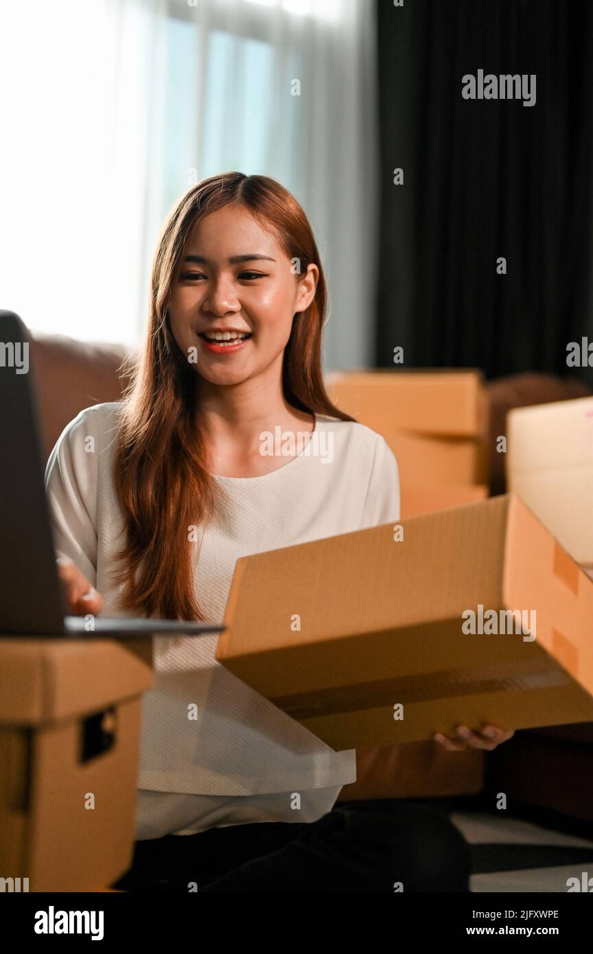 Portrait of a beautiful millennial asian female SME business owner updating her stock and shipping status on laptop computer while preparing a shippin Stock Photo
