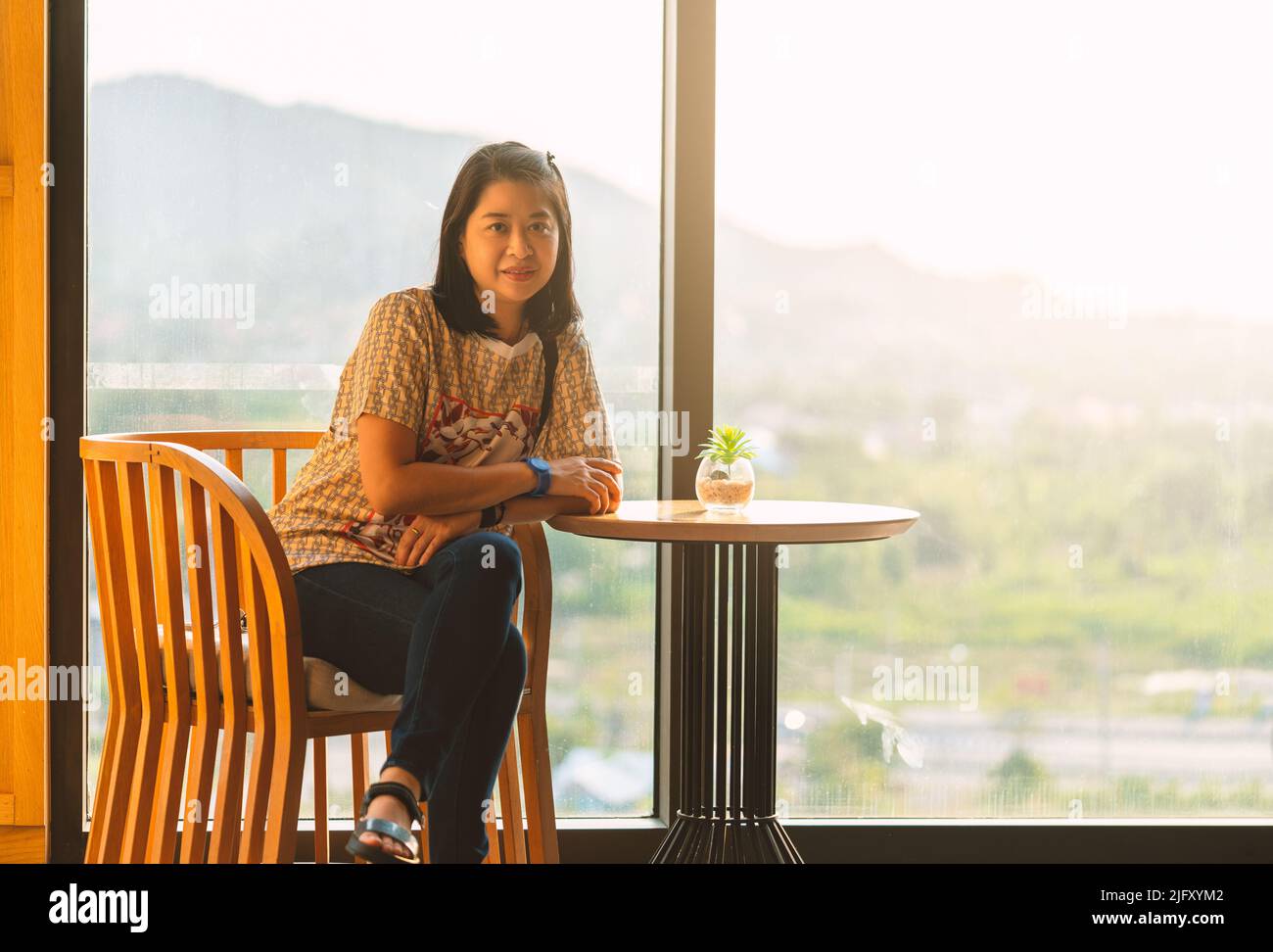 Portrait of Asian middle-aged woman sit on chair, arm on small desk with pot plant, sitting in front of two big mirror window, sunlight from behind. H Stock Photo