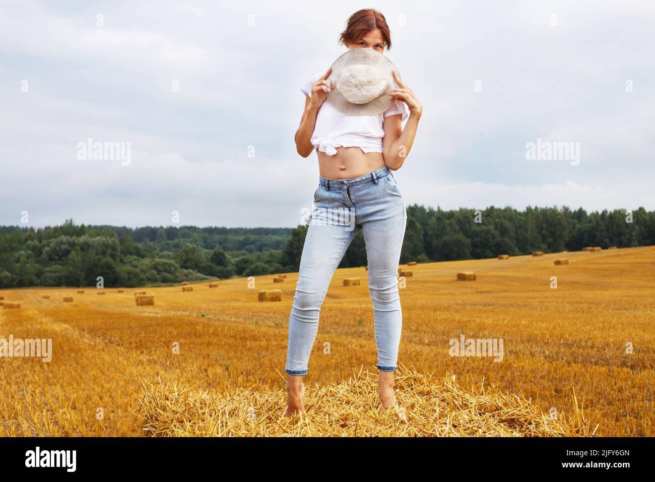 barefoot girl in straw hat stands on a haystack on a bale in the agricultural field after harvesting. Stock Photo