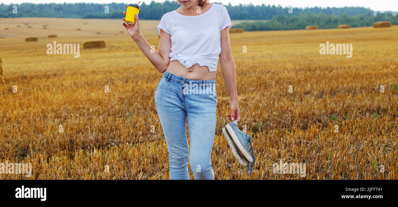 barefoot girl with sneakers and cardboard cup with coffee in hand standing in the agricultural field with haystack and bales after harvesting. Stock Photo