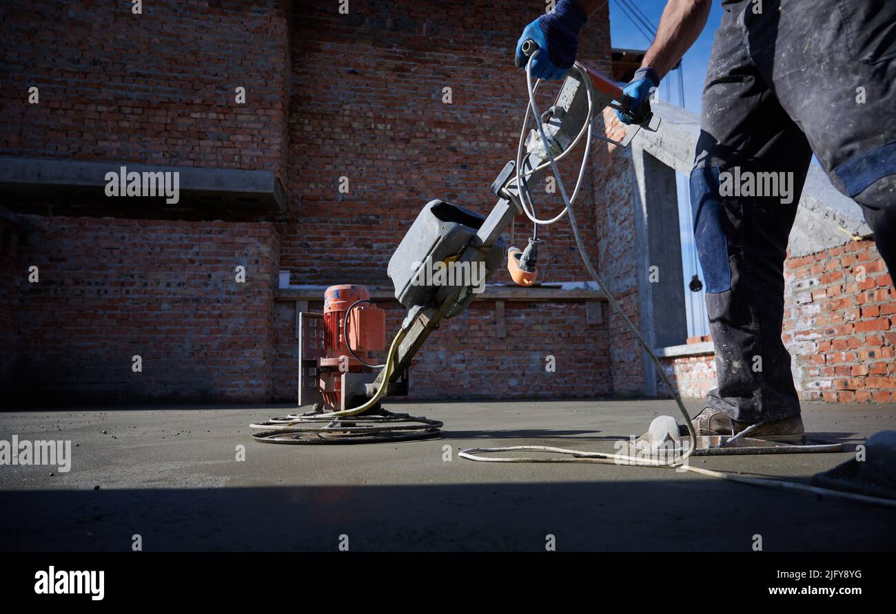 Close up male builder hands in protective gloves holding grout-grinding machine on concrete surface on the background of brick wall outside. Stock Photo