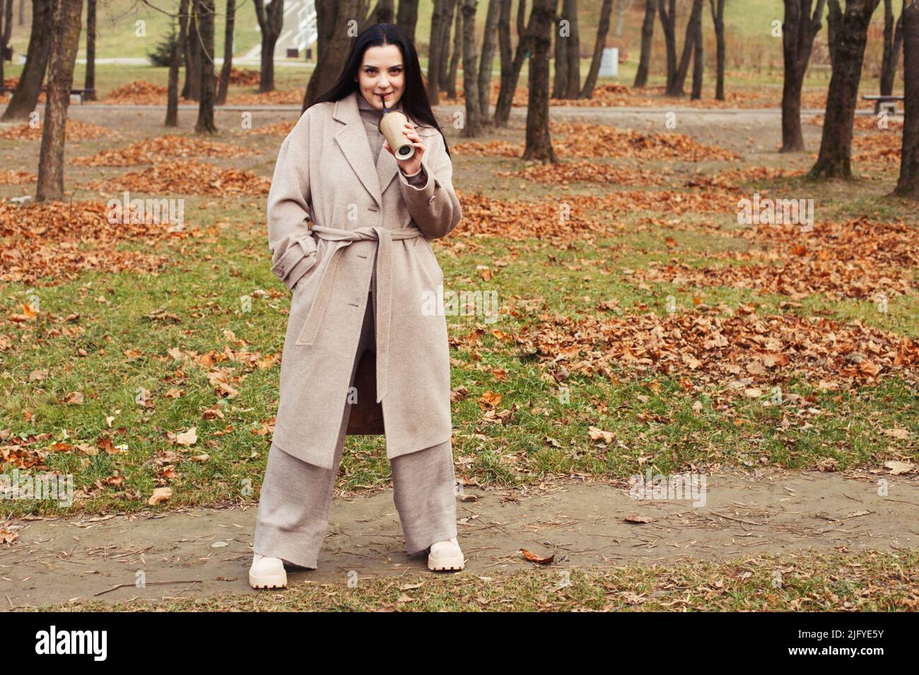 woman in a gray coat walking in the autumn park with a cup of hot drink. Stock Photo