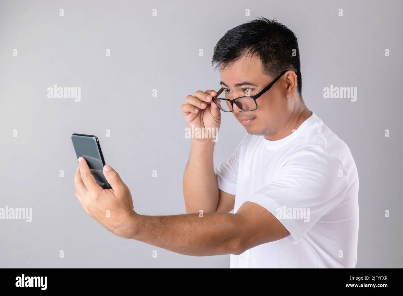 Short or long sighted concept : Man weraing eyeglasses and trying to look clearly on smarthphone in studio shot on grey background Stock Photo