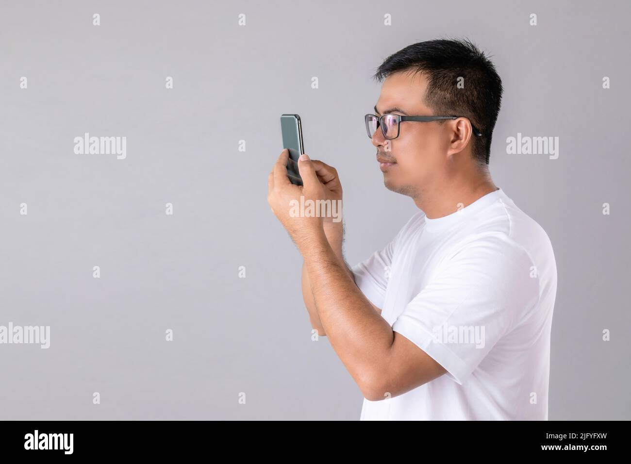 Short or long sighted concept : Man weraing eyeglasses and trying to look clearly on smarthphone in studio shot on grey background Stock Photo