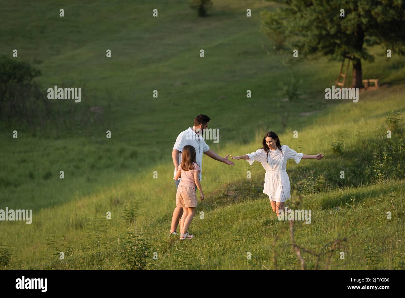 woman extending hand while running on grassy slope towards family Stock Photo