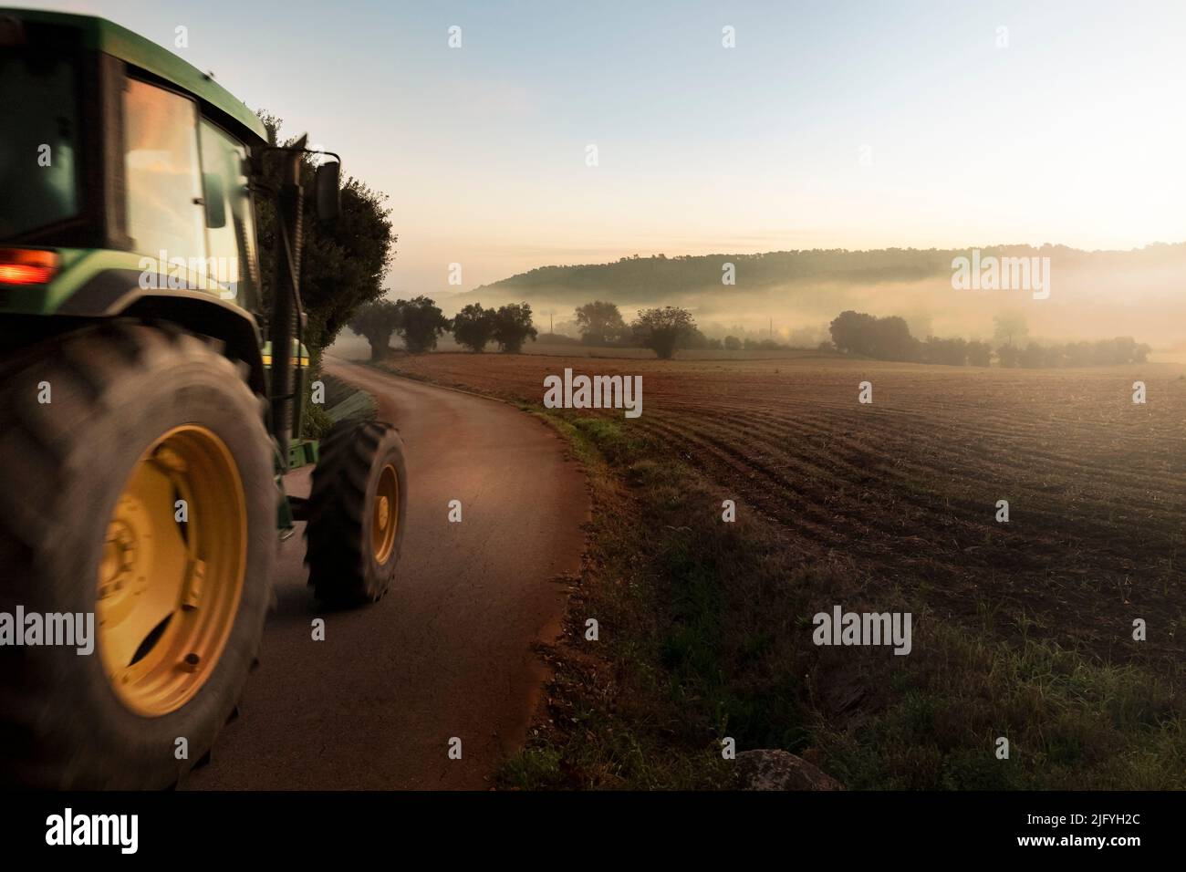 Field tractor riding through farm road at dawn Stock Photo