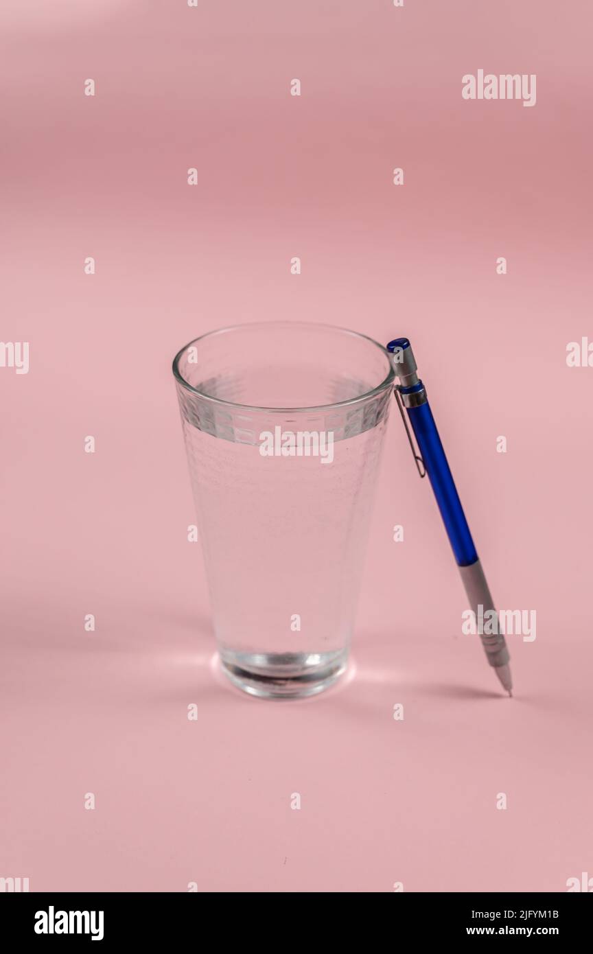 A glass of water and a pencil on a pink background. A full glass of