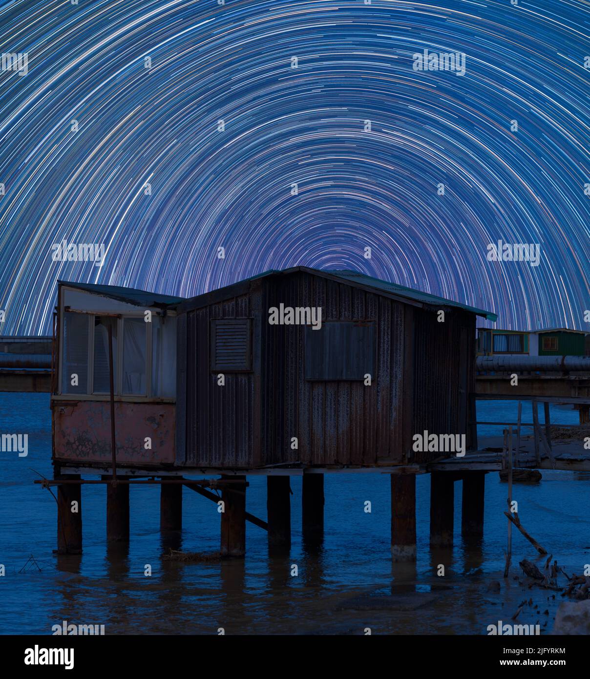 Star trails in the night sky in Italy. Movement of the stars behind an old metal and wooden fishing shed standing in the water Stock Photo