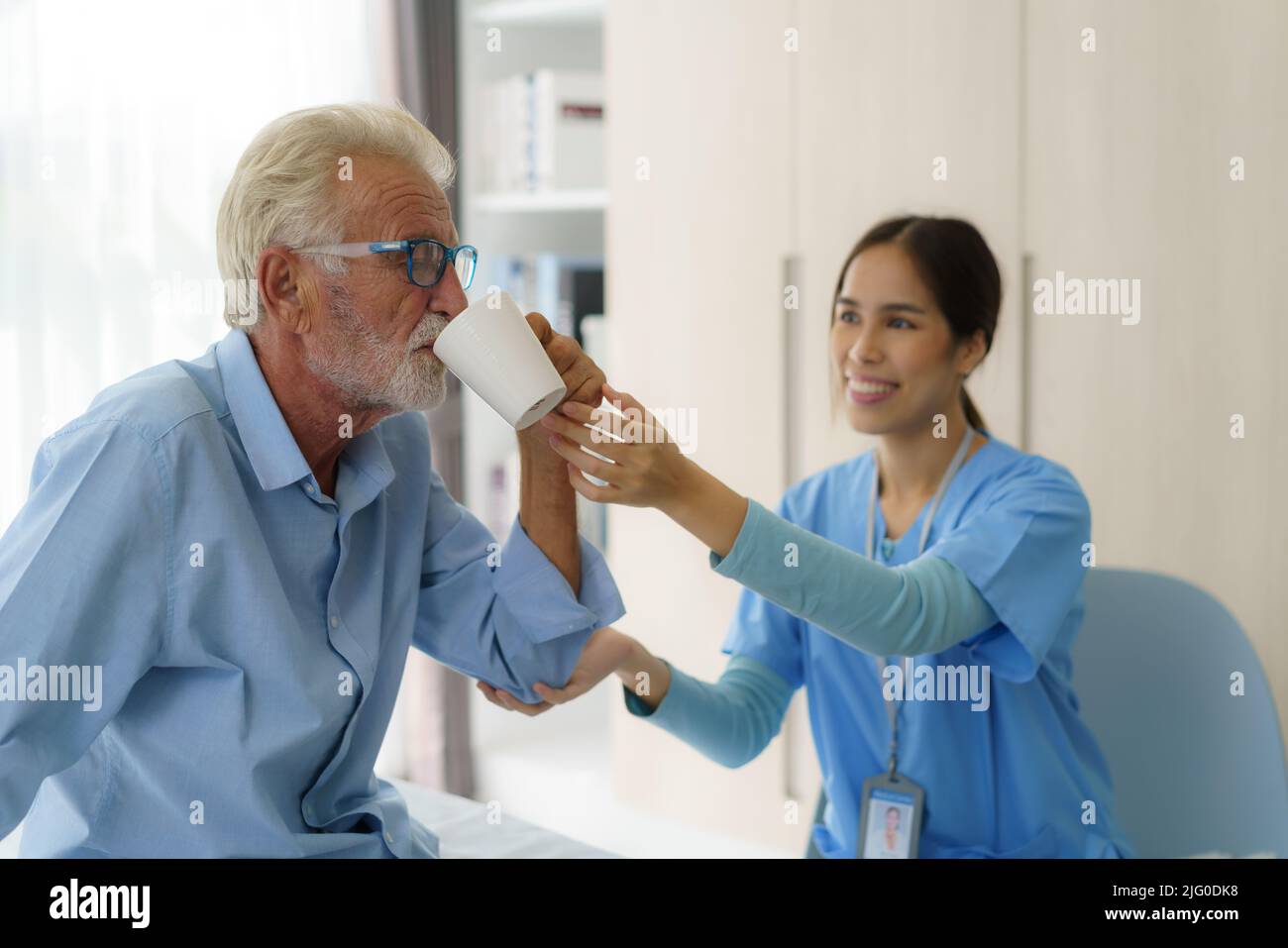 Asian elderly caregiver woman sitting on a hospital bed next to an older man helping drink a glass of water from the bedroom at home. Stock Photo