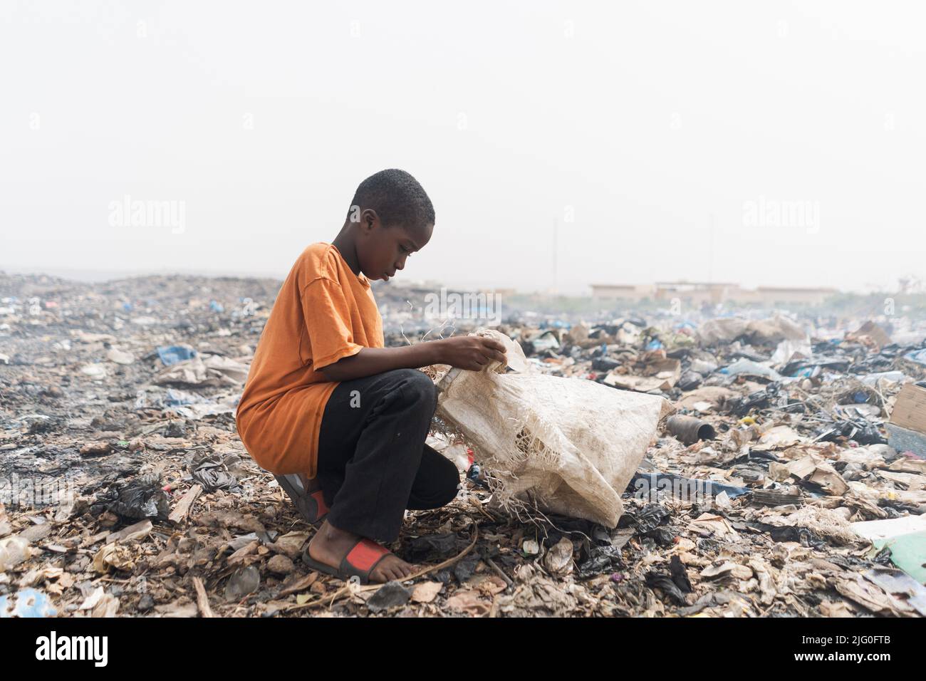 Young African boy collecting recyclable items in a large plastic bag from heaps of rubbish piled up in a smelly landfill Stock Photo