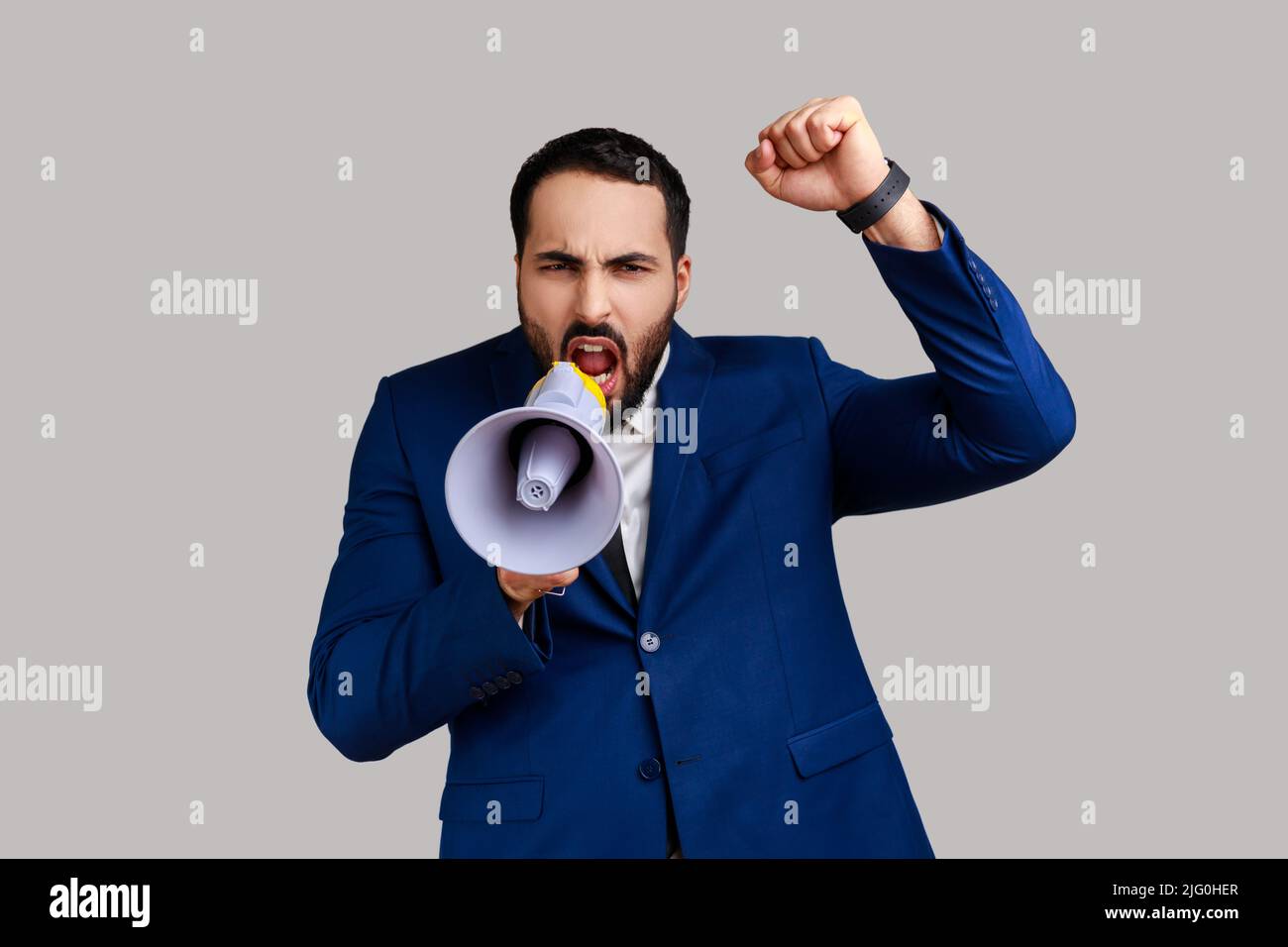 Portrait of bearded businessman standing with raised hands and holding megaphone, screaming in loud speaker, protesting, wearing official style suit. Indoor studio shot isolated on gray background. Stock Photo