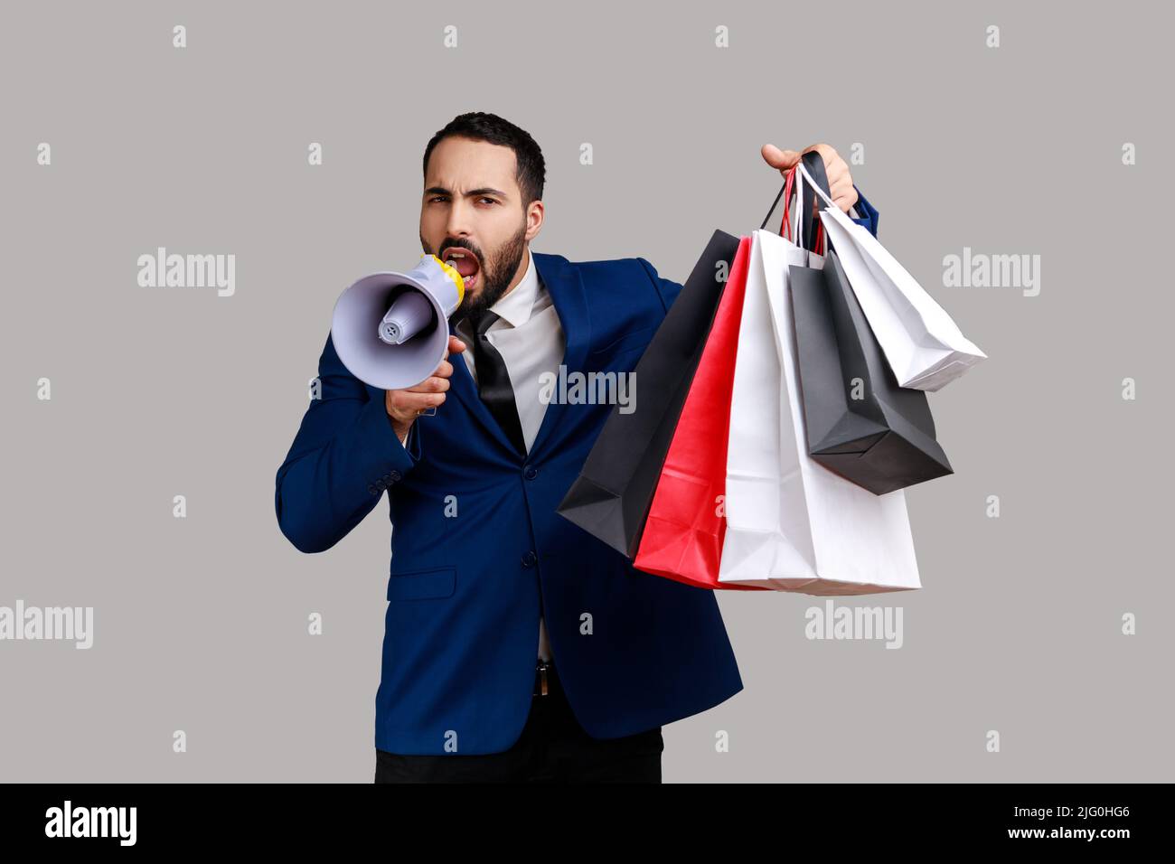 Bearded man announcing shopping discounts and sale, holding shopping bags with purchases, screams in loud speaker, wearing official style suit. Indoor studio shot isolated on gray background. Stock Photo