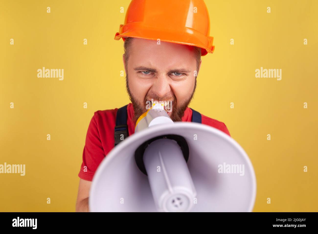 Portrait of angry aggressive worker man wearing uniform and protective helmet taking selfie, point of view of photo, screaming in megaphone. Indoor studio shot isolated on yellow background. Stock Photo