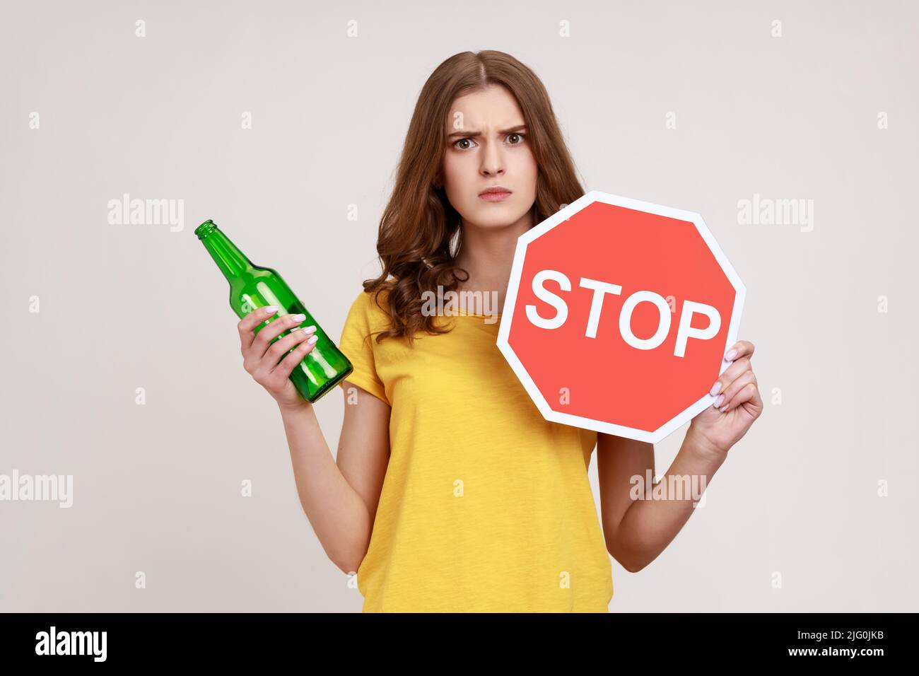 Stop drinking alcohol. Serious teenager girl in yellow T-shirt holding red stop sign and alcohol bottle, looking at camera strict expression. Indoor studio shot isolated on gray background. Stock Photo