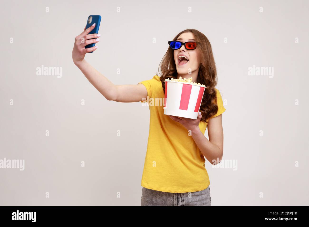 Young funny happy excited teenager girl wearing yellow casual style T-shirt holding takeaway popcorn bucket, making selfie or broadcasting livestream. Indoor studio shot isolated on gray background. Stock Photo
