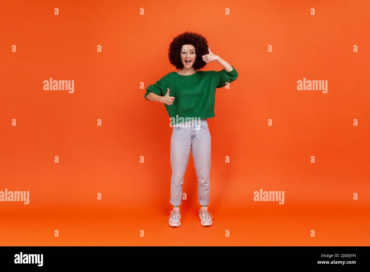 Full length portrait of happy friendly woman with Afro hairstyle wearing green casual style sweater standing with thumbs up, expressing positive. Indoor studio shot isolated on orange background. Stock Photo