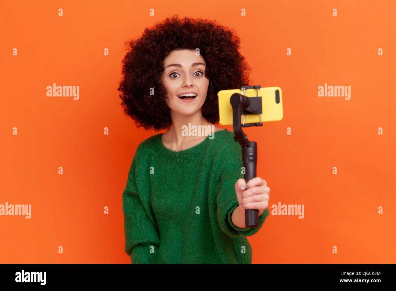 Portrait of woman blogger with Afro hairstyle wearing green casual style sweater using phone and steadicam, broadcasting livestream. Indoor studio shot isolated on orange background. Stock Photo